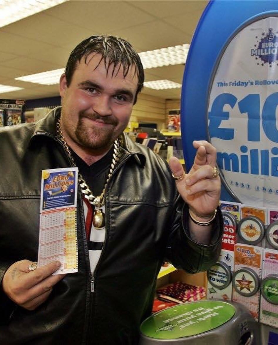 A man in a leather jacket holding a EuroMillions lottery ticket smiles and gestures for luck next to a sign displaying a £10 million jackpot.