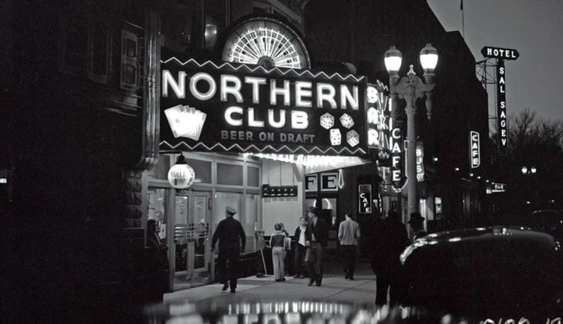 Black and white photo of people outside the brightly lit Northern Club bar at night, with neon signs reading “BEER ON DRAFT” and nearby signs for a hotel, café, and bar visible along the street.