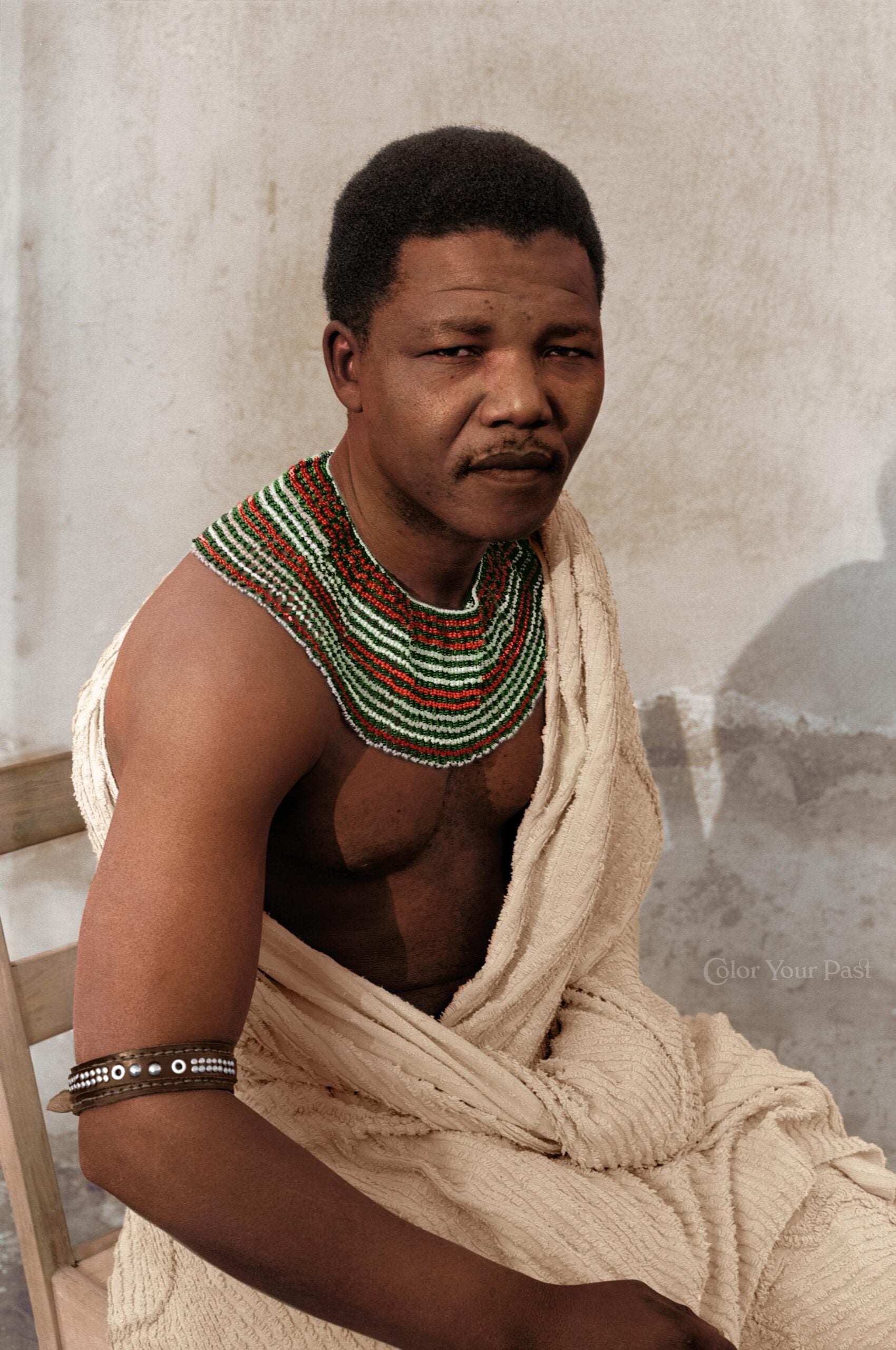 A man with short hair wears a cream-colored draped garment, a colorful beaded necklace, and an armband. He sits on a wooden chair against a plain, light-colored wall, looking slightly to the side.