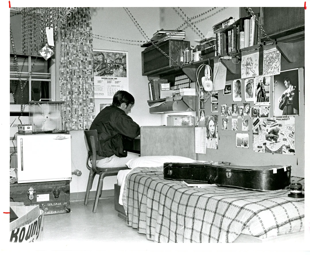 A person sits at a desk in a cluttered dorm room with posters, books, a guitar case on a bed, a small fridge, and various items on shelves and walls.