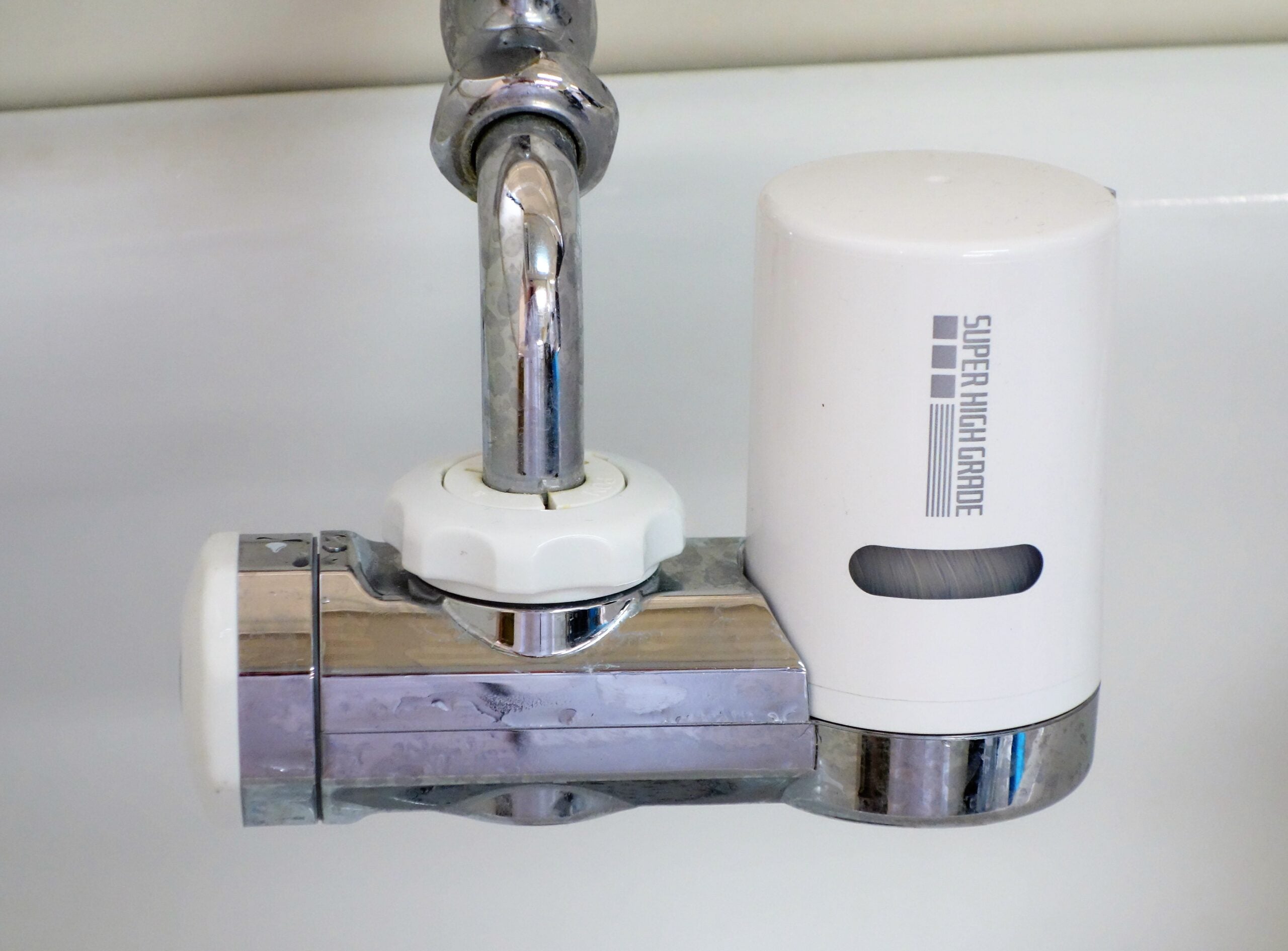A close-up of a chrome faucet with a white water filter attachment labeled “SUPER HIGH GRADE” installed on it. The background is a white sink.