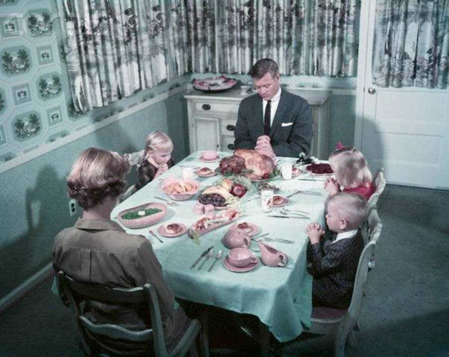 A family of five sits around a dining table, heads bowed in prayer before a meal. The table is set with food and dishes, and the room is decorated in pastel colors with patterned wallpaper and curtains.