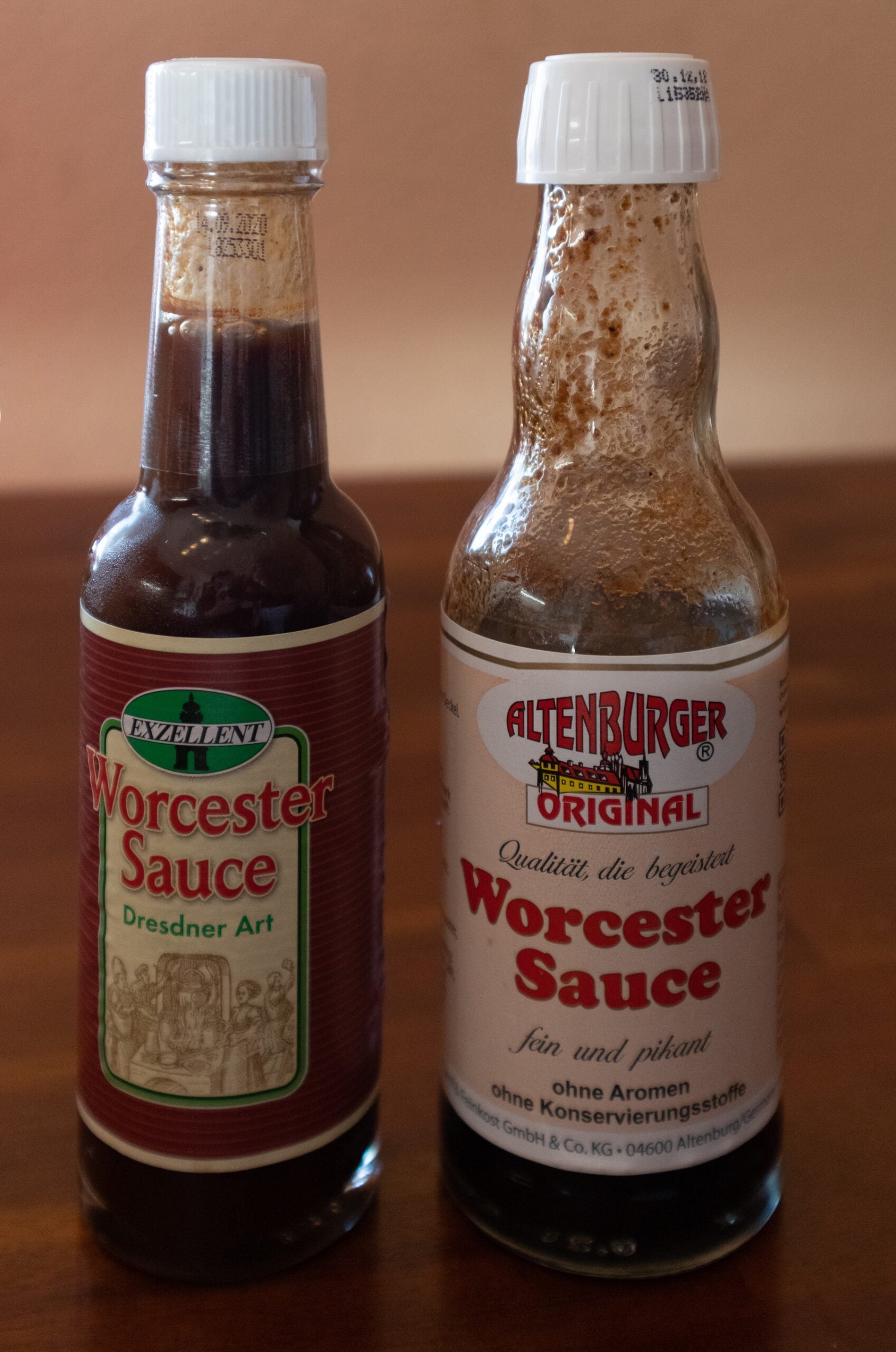 Two bottles of Worcester sauce on a wooden surface; the bottle on the left has a dark red label, and the bottle on the right has a white label with sauce splashes near its neck.