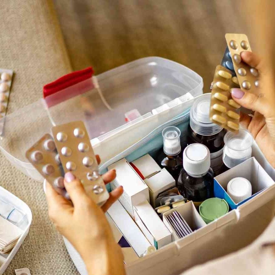 A person holds blister packs of pills above an open first aid box filled with various medicines, bottles, and medical supplies.