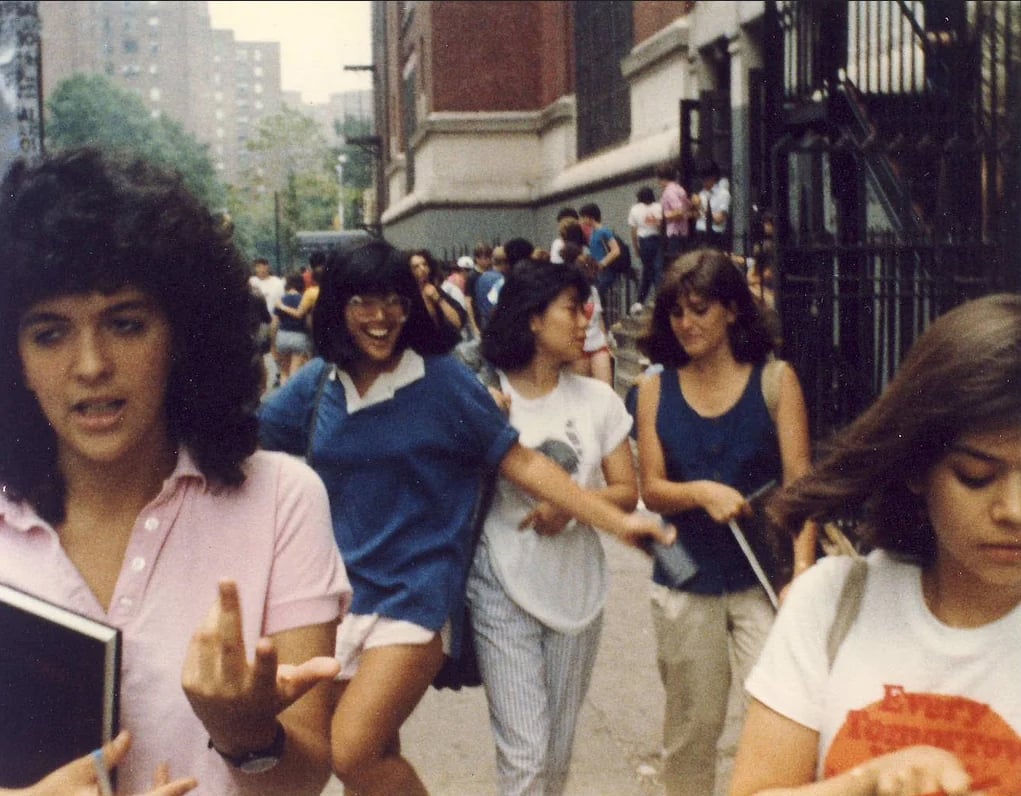A group of young people walk outside near a school. Some are smiling, holding books, and chatting. The background shows a city street and buildings. One person in front gestures with their hand.
