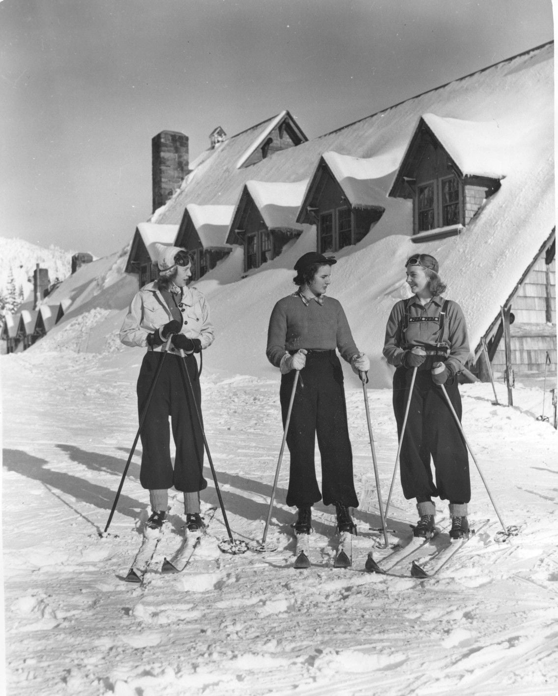 Three women in vintage winter clothing stand with skis and poles on snow in front of a row of snow-covered buildings, smiling and conversing.