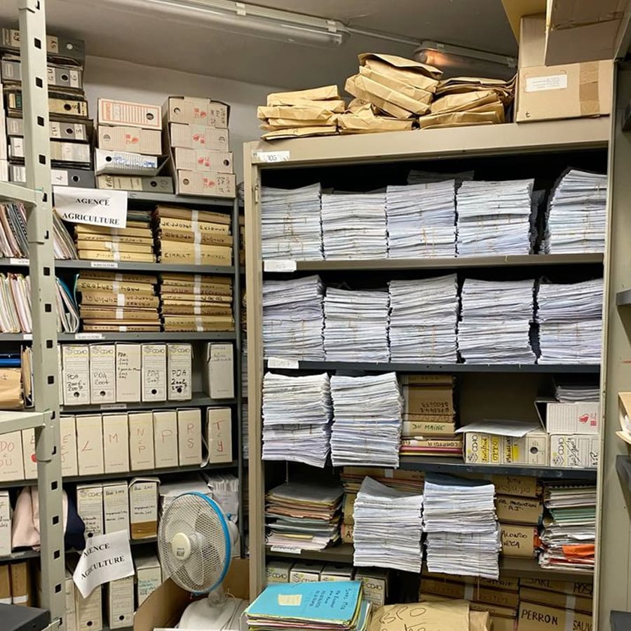 A cluttered archive room with shelves full of stacked papers, folders, labeled boxes, and bundles of documents. A small electric fan is on the floor among files and boxes.