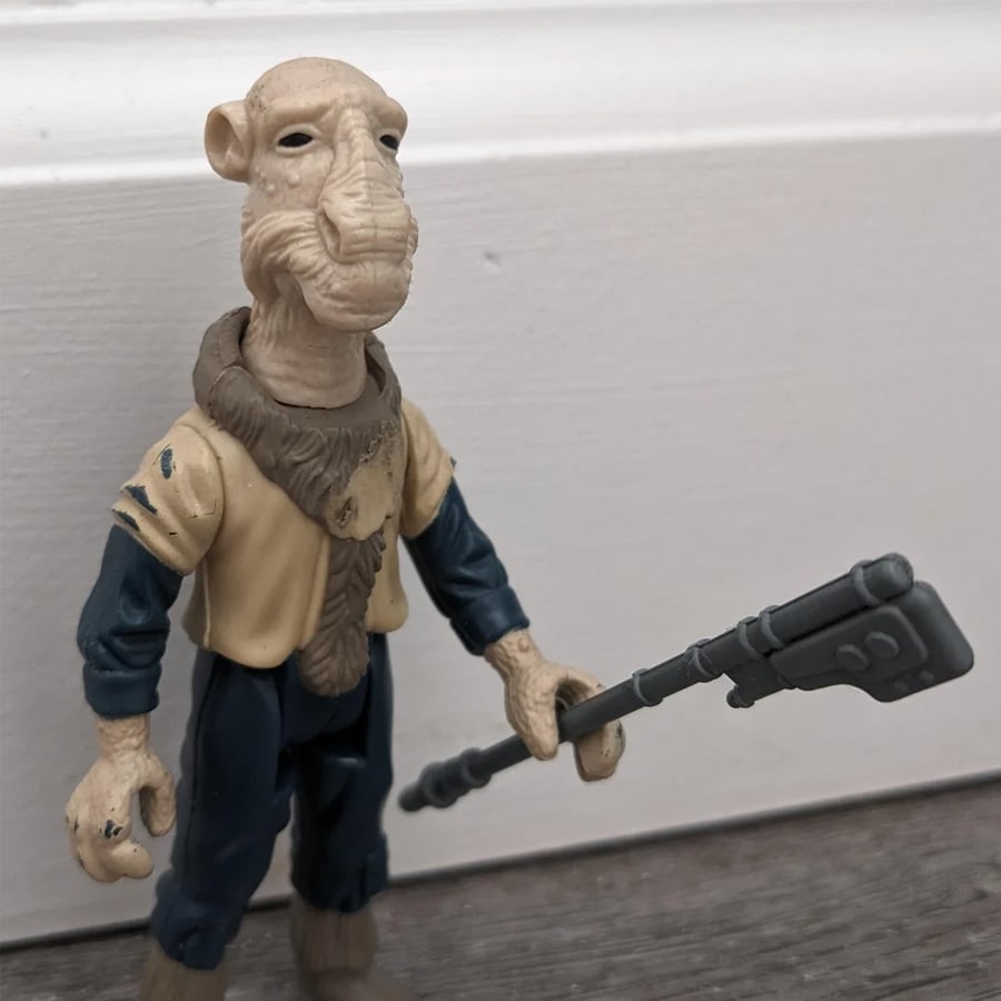 A beige alien action figure with a textured head, wearing a tan top and dark pants, holds a long, gray staff. The figure stands on a wooden floor against a white wall.