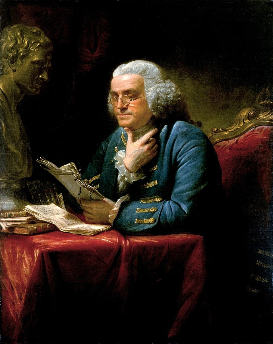 An older man with white hair and glasses sits at a red-draped table, holding papers and touching his neck, with books and a statue beside him, in a dimly lit room.