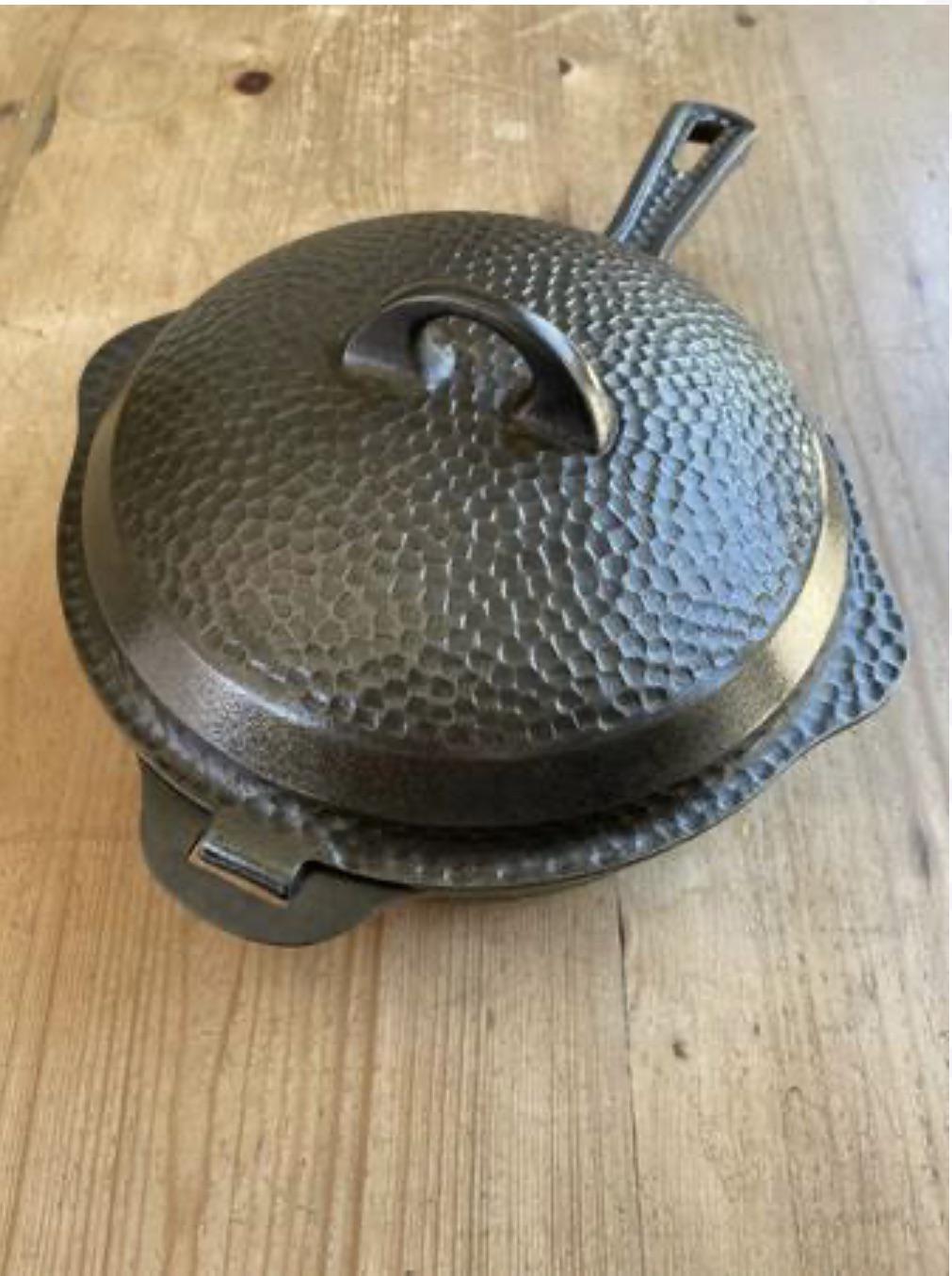 A textured, cast iron Dutch oven with a lid sits on a wooden surface. The pot has a hammered design and two handles, one on each side.