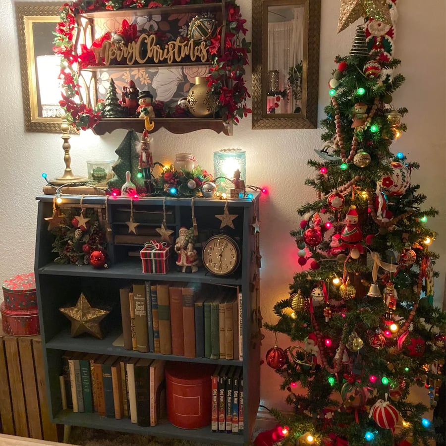 A decorated Christmas tree with colorful lights stands next to a blue bookshelf filled with books, ornaments, wrapped gifts, and a clock. A “Merry Christmas” sign, wreath, and lights add festive cheer to the cozy room.