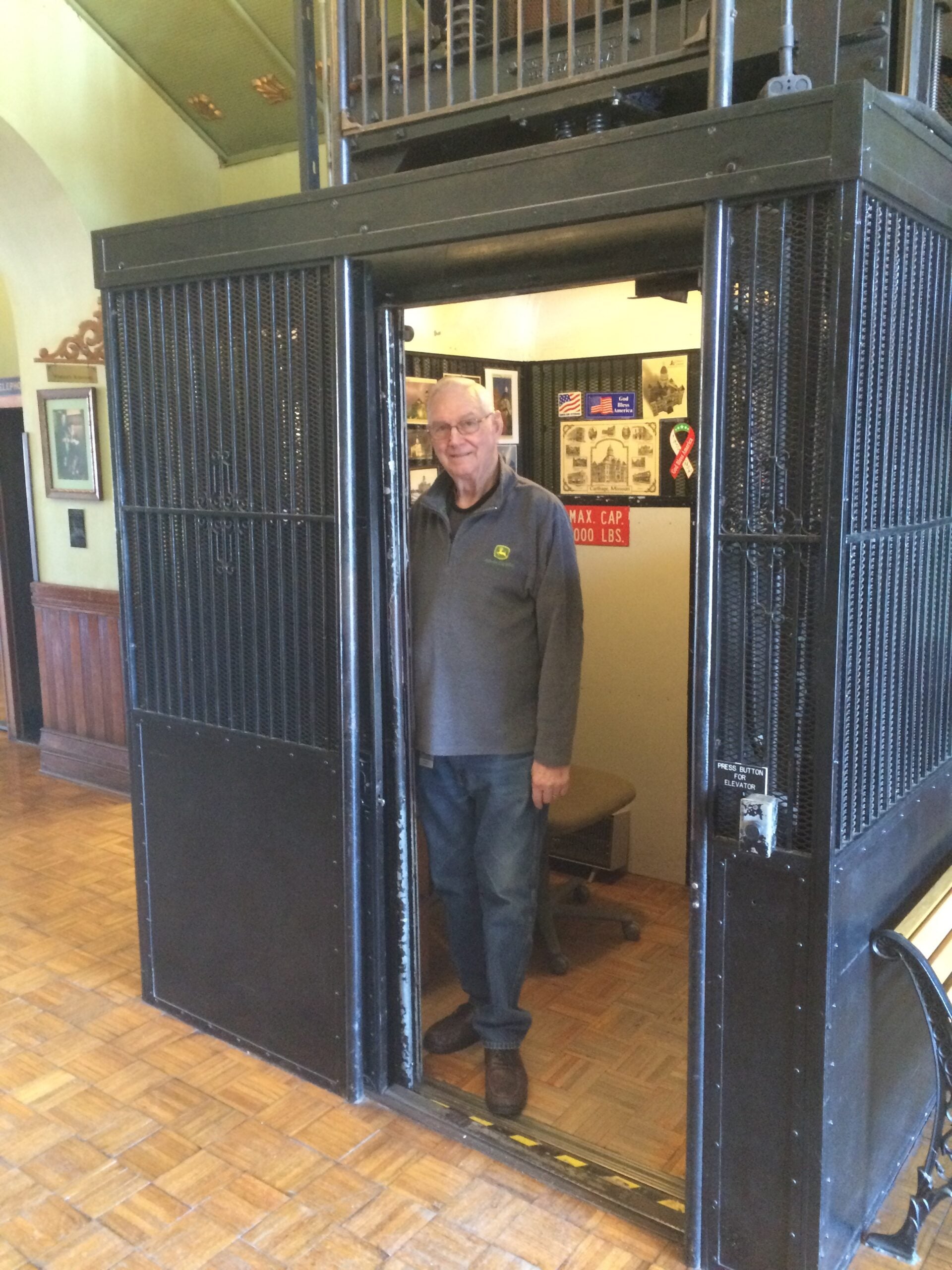 An older man stands inside an open, vintage black elevator with metal gates in a wood-paneled room. Posters and signs are visible inside the elevator behind him.