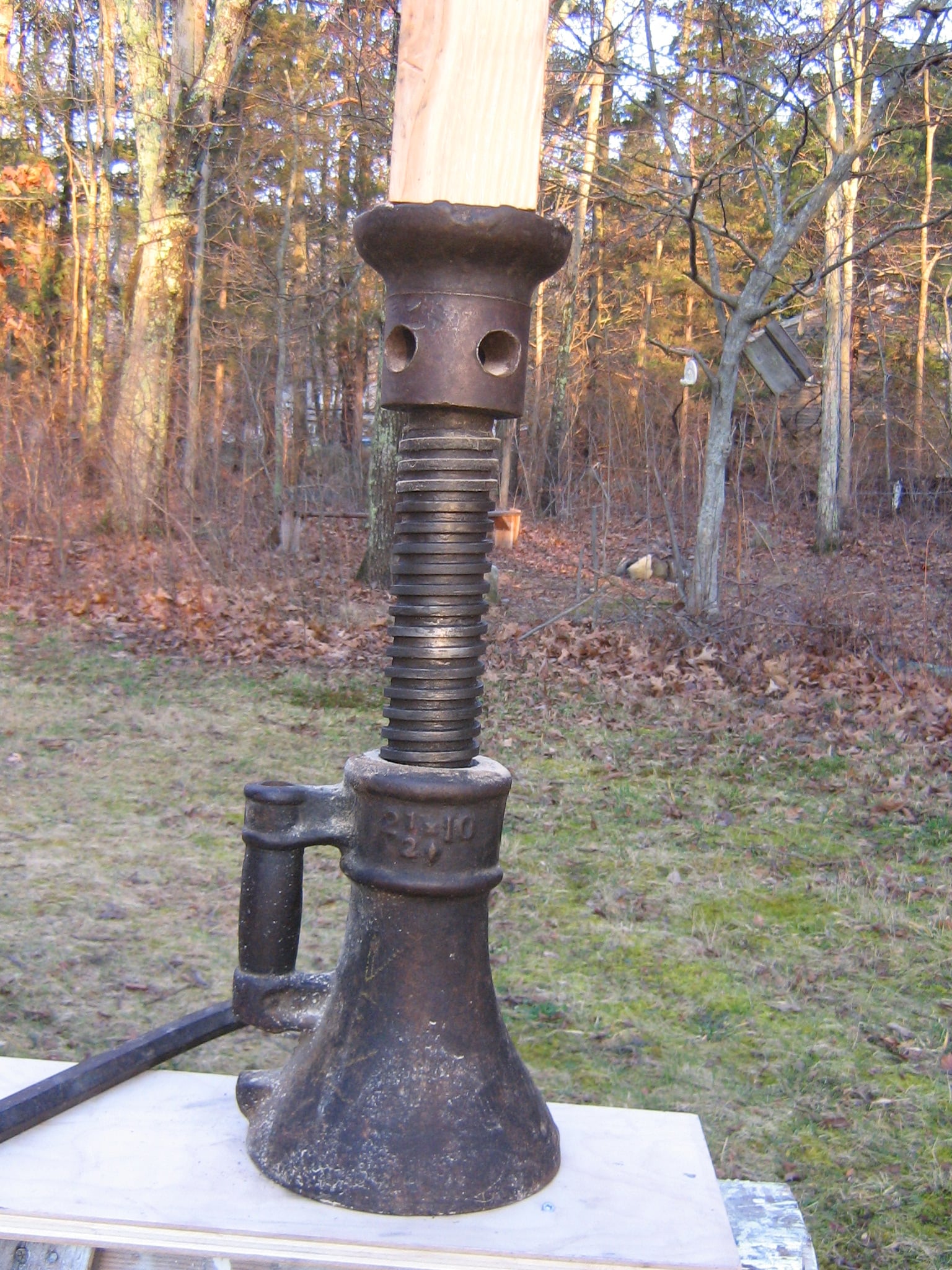 A vintage mechanical jack with a large threaded rod and a handle stands outdoors on a wooden surface, surrounded by trees and dry leaves.