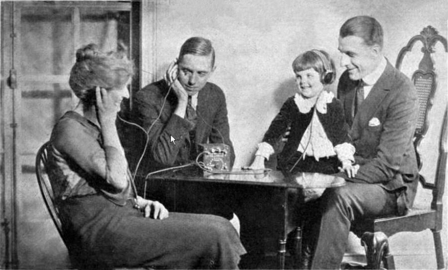 A woman, two men, and a young girl sit around a table, each listening with headphones. The girl smiles, and a vintage radio device sits on the table.