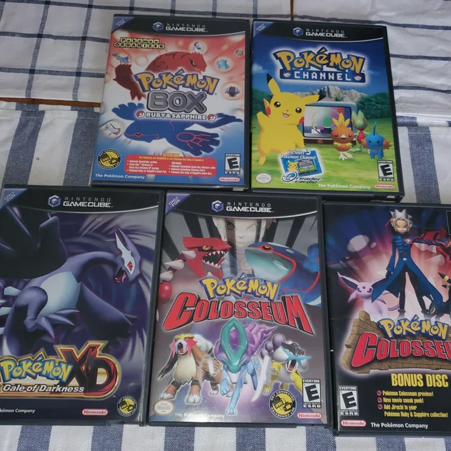 Five Nintendo GameCube Pokémon game cases are arranged on a checkered cloth: Pokémon Box, Pokémon Channel, Pokémon XD: Gale of Darkness, Pokémon Colosseum, and Pokémon Colosseum Bonus Disc.