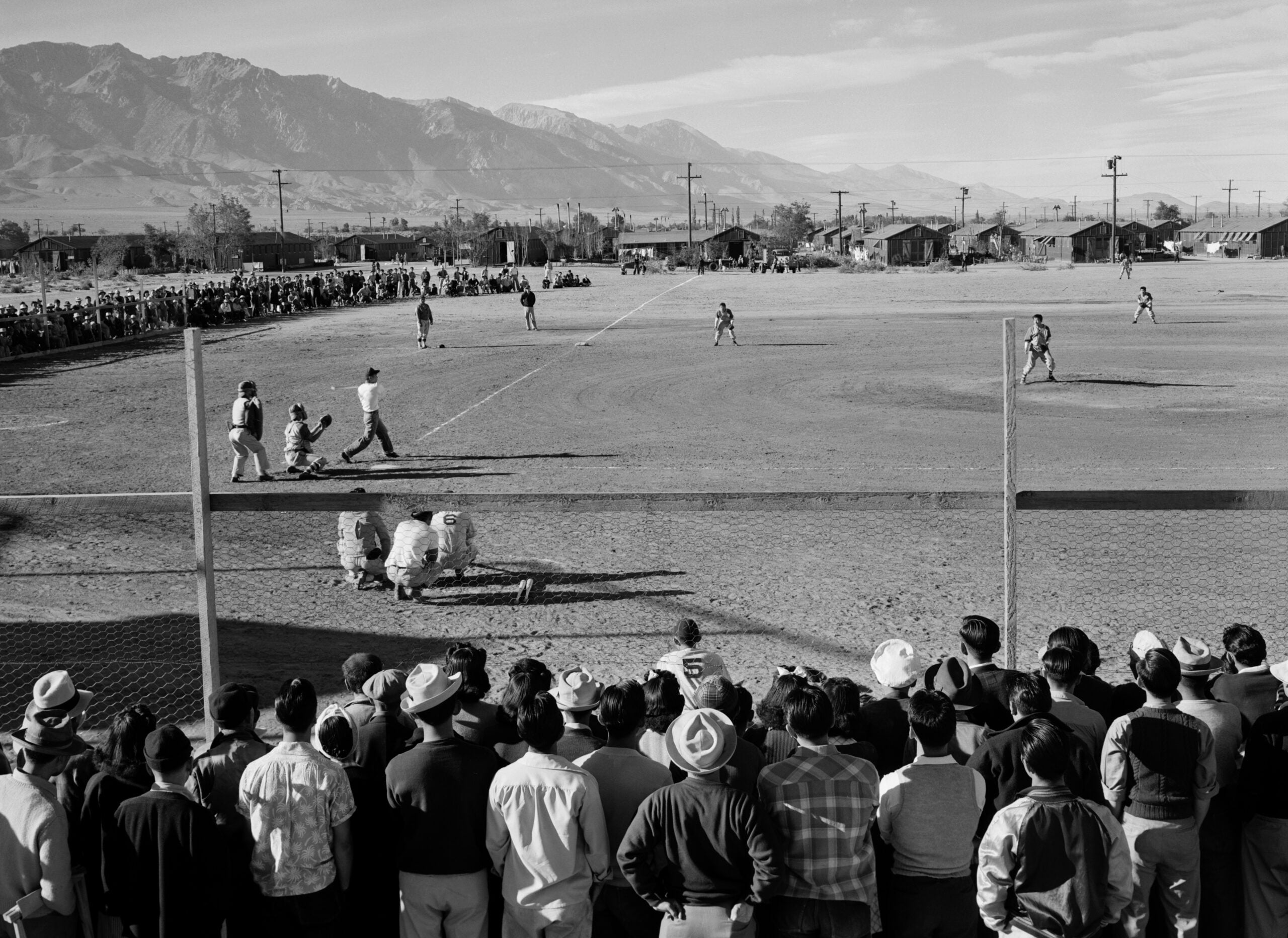 A crowd watches a baseball game on a dirt field with mountains in the background and rows of small buildings nearby, under a clear sky.