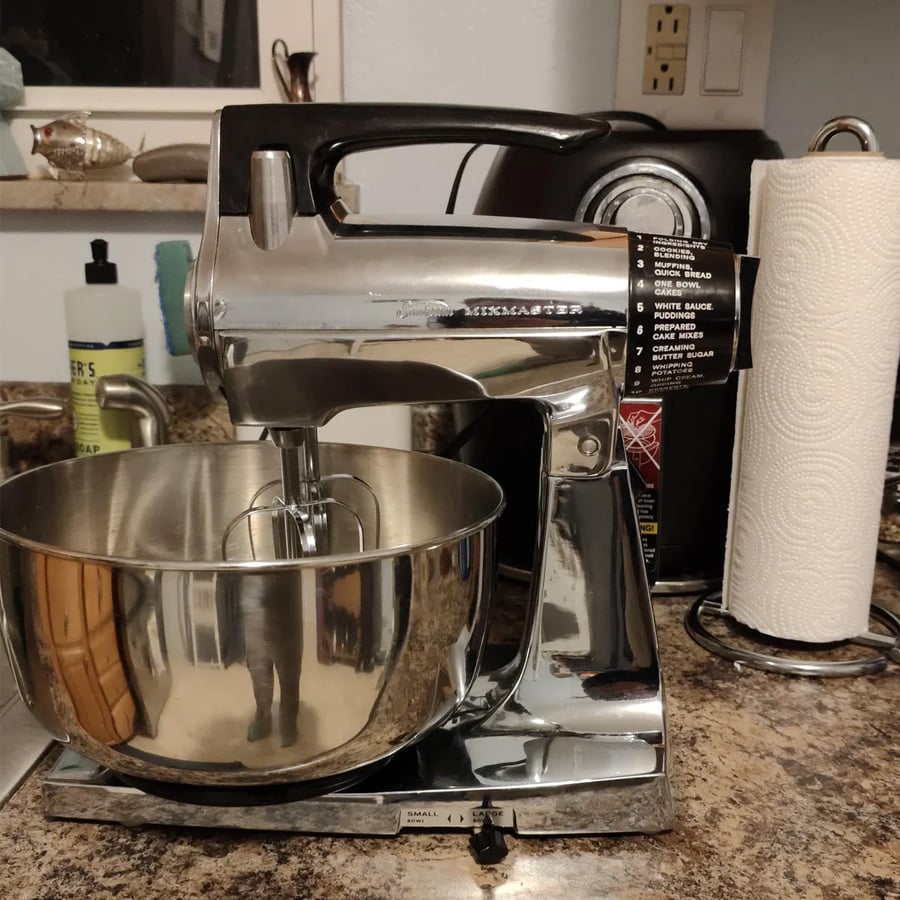 A vintage silver stand mixer with a stainless steel bowl sits on a kitchen counter next to a paper towel roll, soap, and other kitchen items.