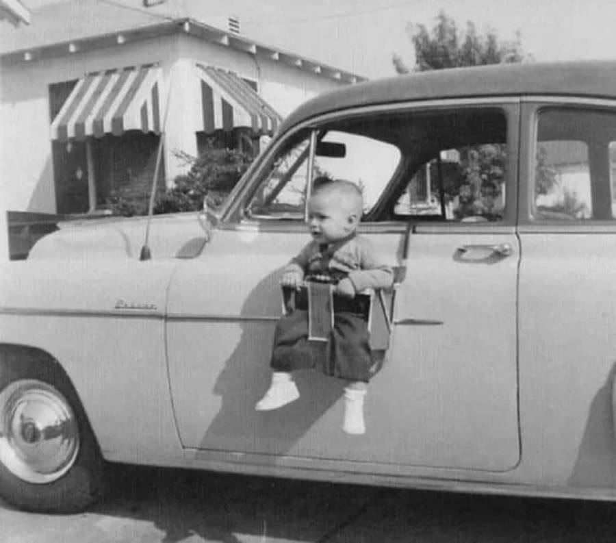 A baby in a harness-style seat is attached to the side of a car door, smiling. The photo is black and white, with a house and striped awning visible in the background.