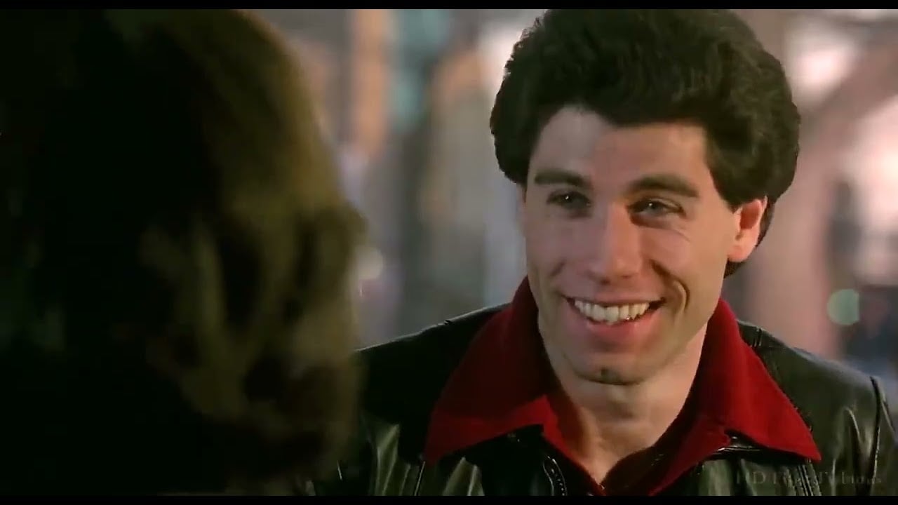 A young man with dark hair smiles while wearing a black leather jacket and red shirt, facing a person with long hair in an outdoor, softly lit setting.