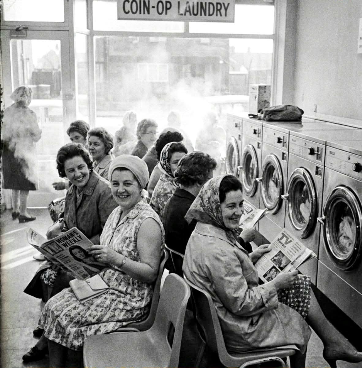 A group of women sit on chairs in a laundromat, some reading magazines, while washing machines run beside them. Sunlight streams in through the window, and a sign above reads “COIN-OP LAUNDRY.”