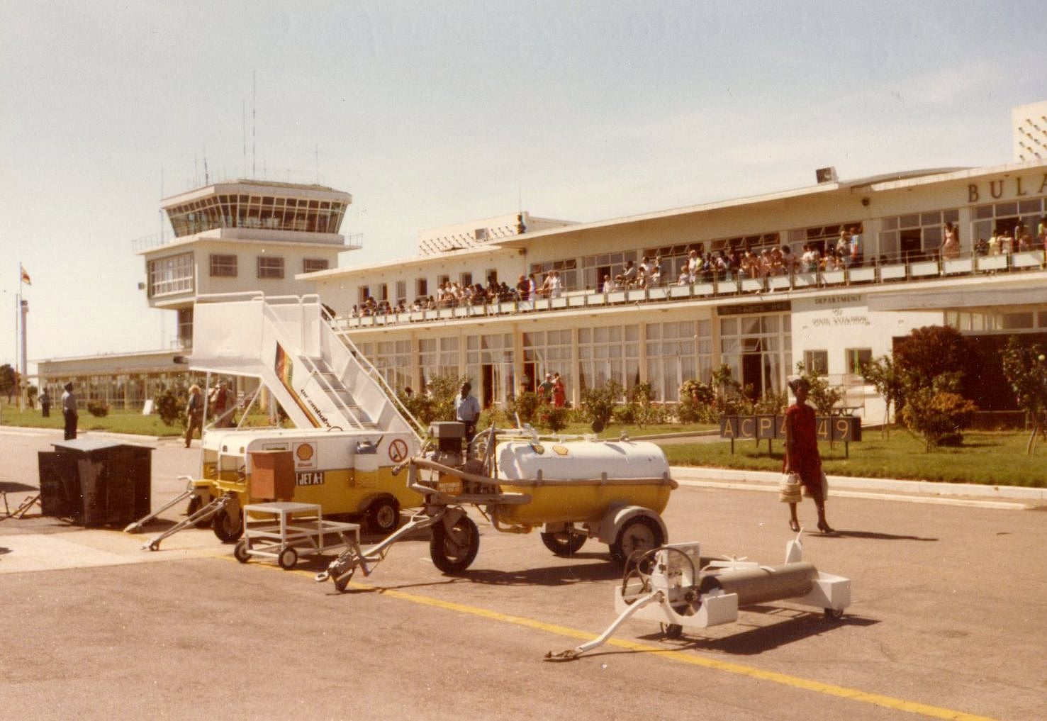 View of an airport terminal with people on a balcony, control tower in the background, and mobile stairs and airport ground equipment in the foreground on a sunny day.