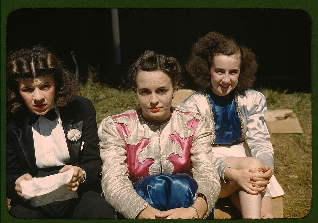 Three young women sit on grass outdoors, wearing vintage clothing. The woman in the center wears a pink and silver jacket, while the others wear dark and light outfits. All look towards the camera, two with neutral expressions, one smiling.