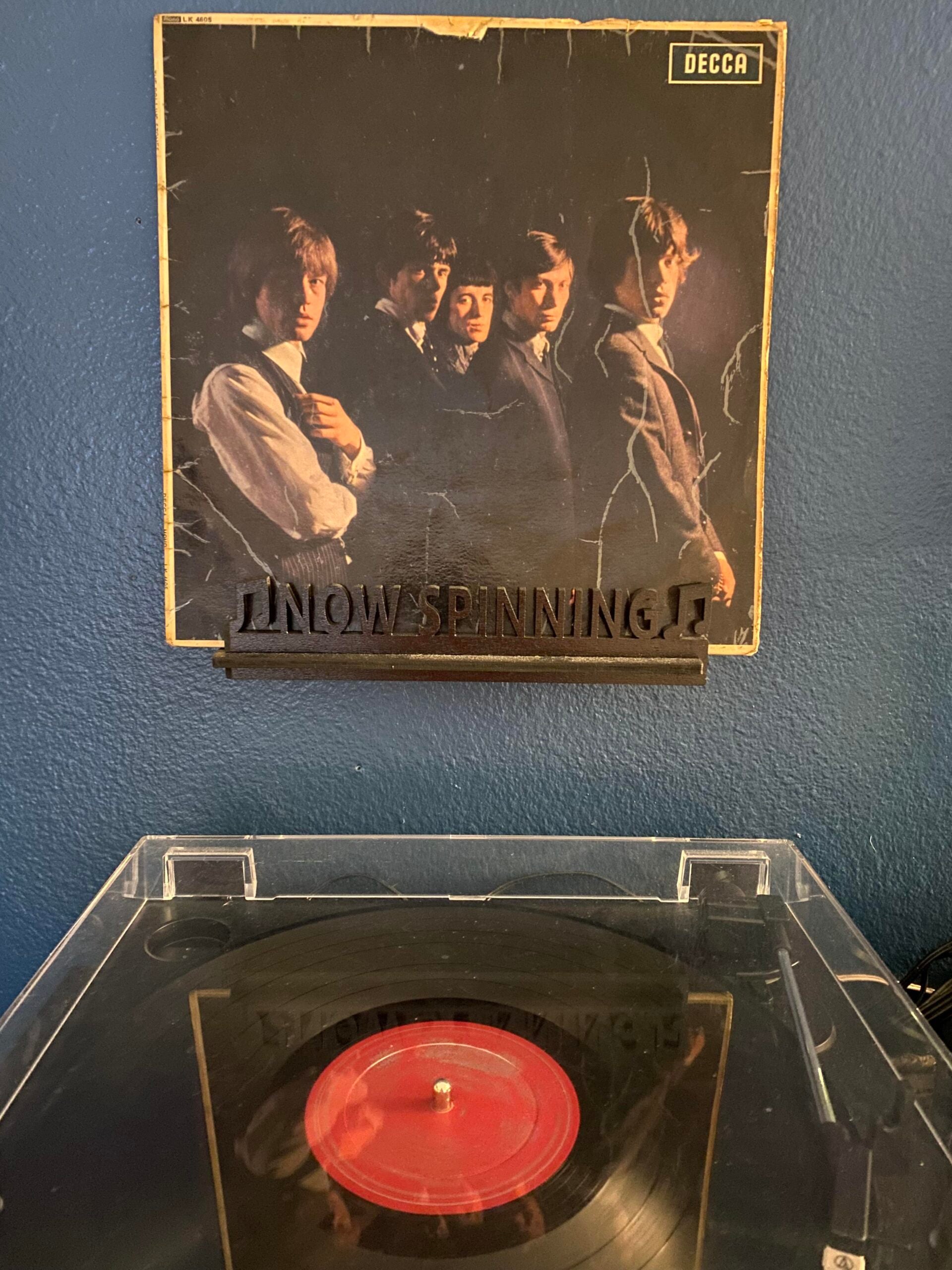 A vinyl record spins on a turntable below a worn album cover of The Rolling Stones displayed on a stand labeled “Now Spinning” against a blue wall.