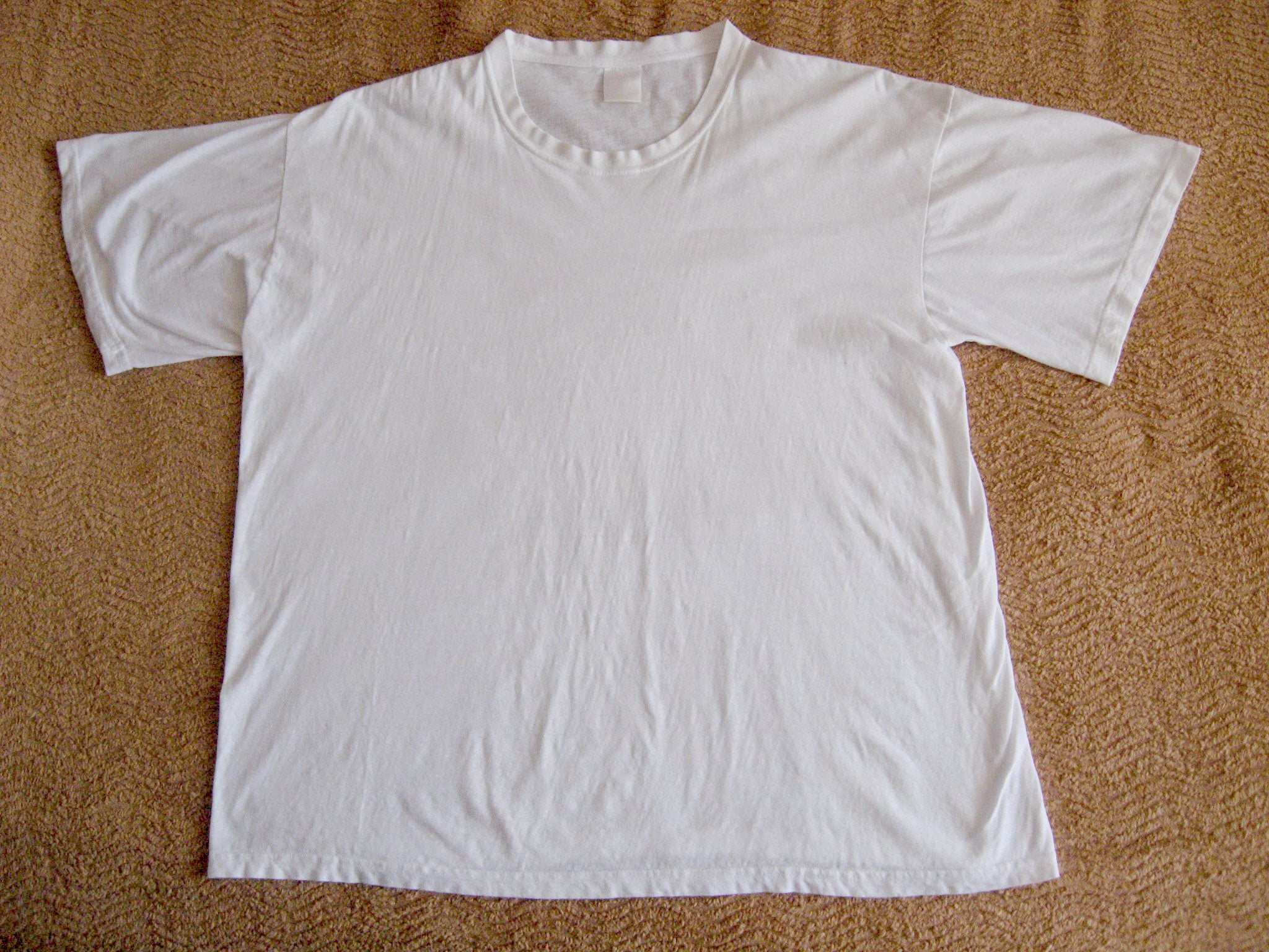 A plain white short-sleeve T-shirt is laid flat on a textured light brown fabric surface.