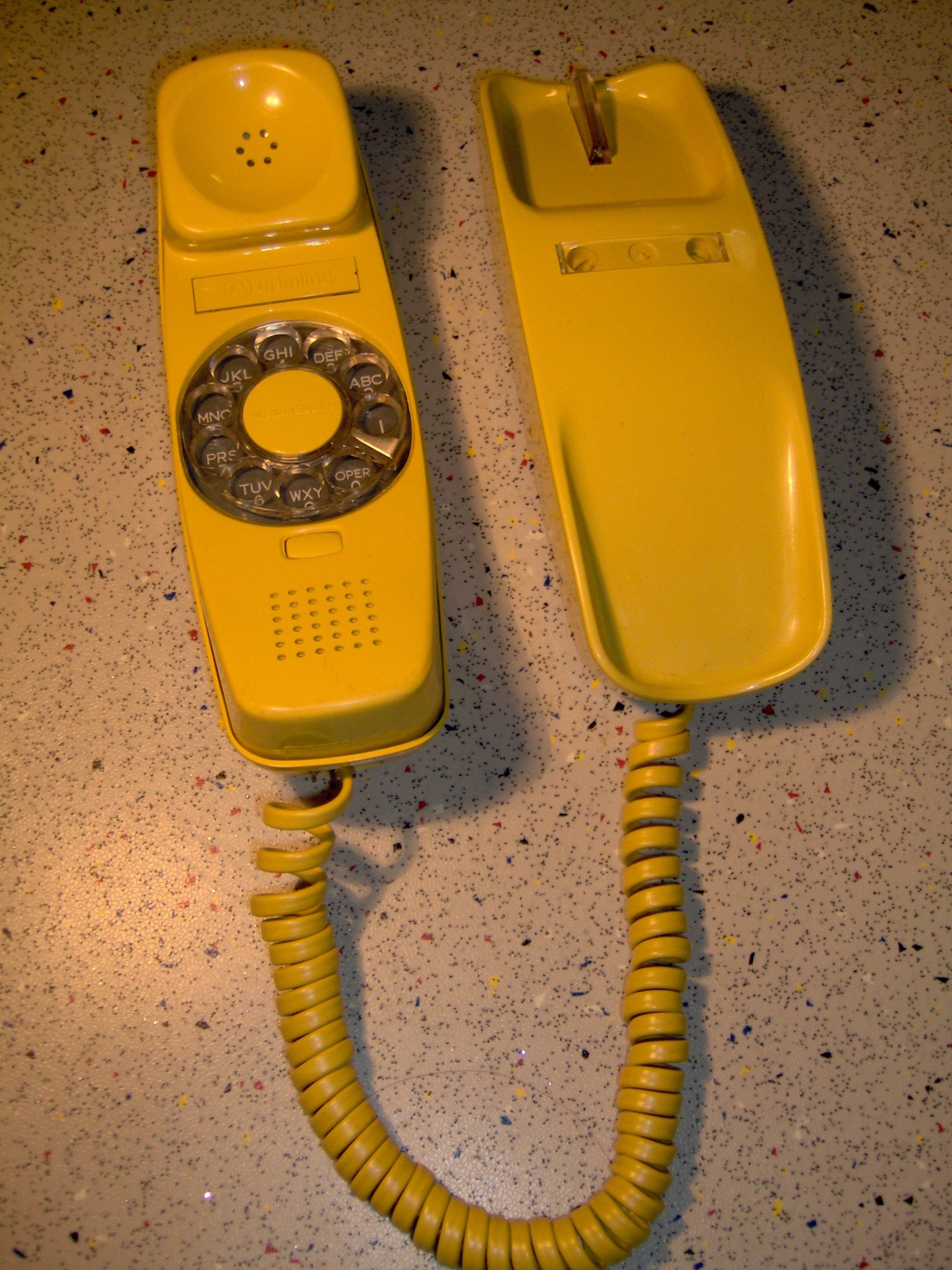 A bright yellow vintage rotary dial telephone with its handset off the hook, connected by a coiled cord, resting on a speckled countertop.