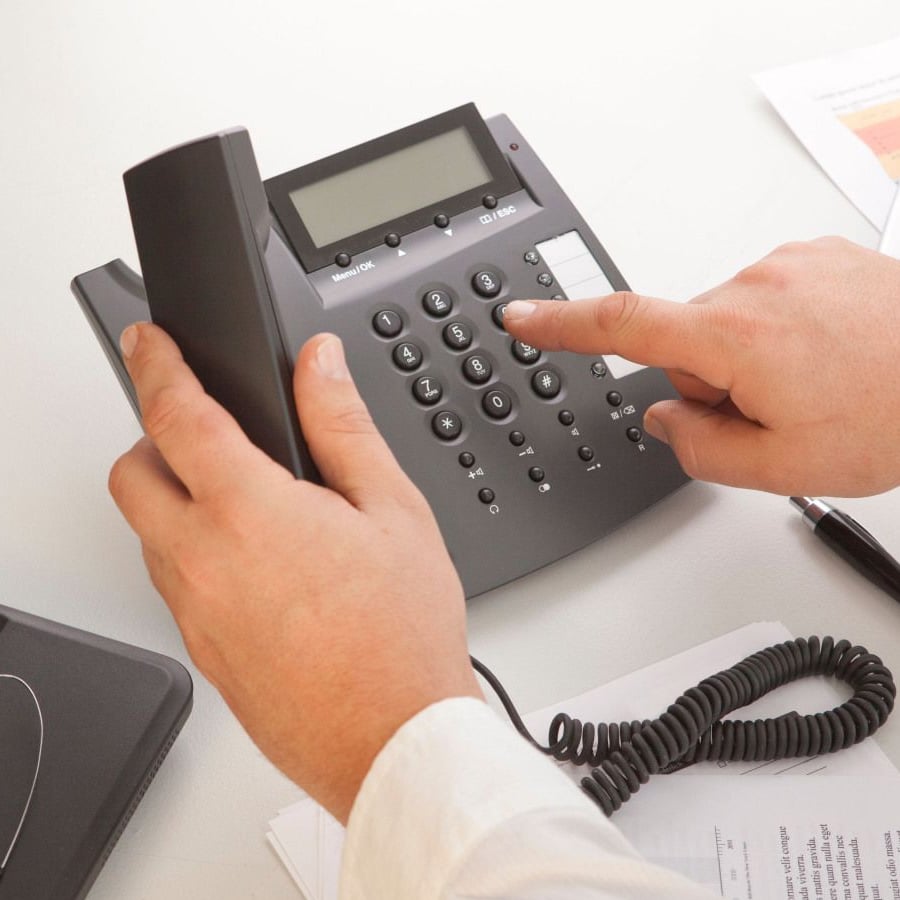A person in a white shirt is dialing a number on an office desk phone, with documents, a pen, and part of a laptop visible on the desk.