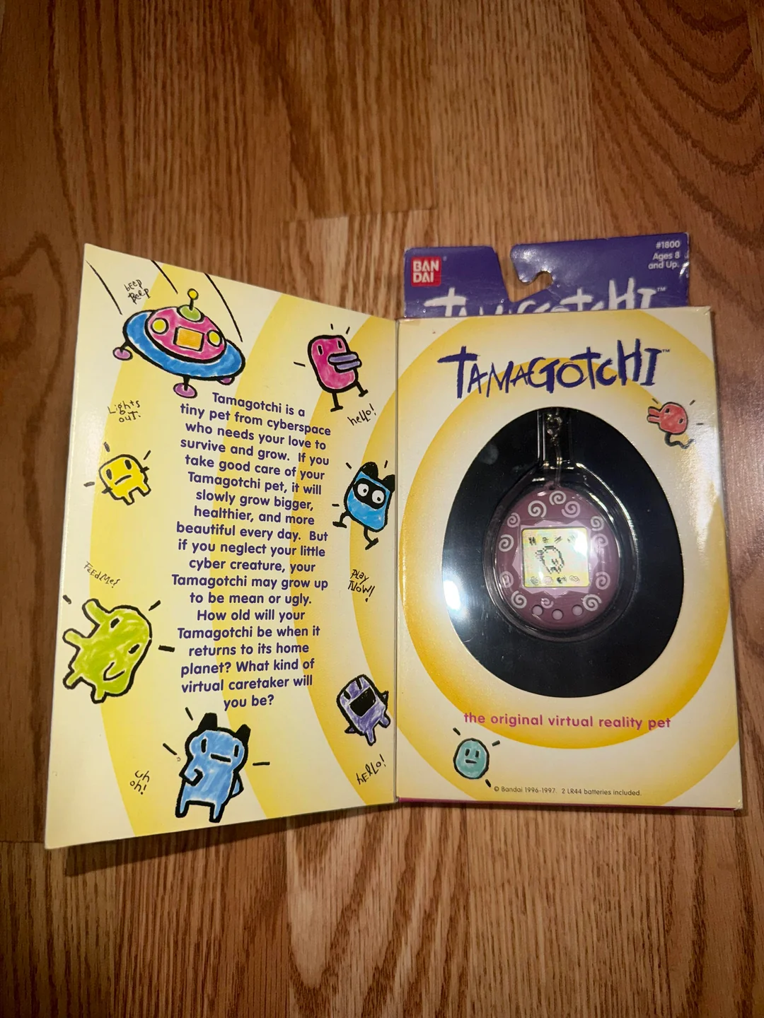 A Tamagotchi virtual pet toy in its original packaging is displayed on a wooden surface. The box is open, showing colorful cartoon characters and playful text explaining the Tamagotchi concept.