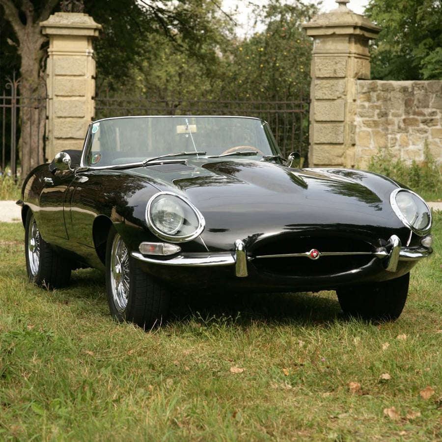 A classic black convertible sports car is parked on grass near a stone wall and gate, with trees in the background.