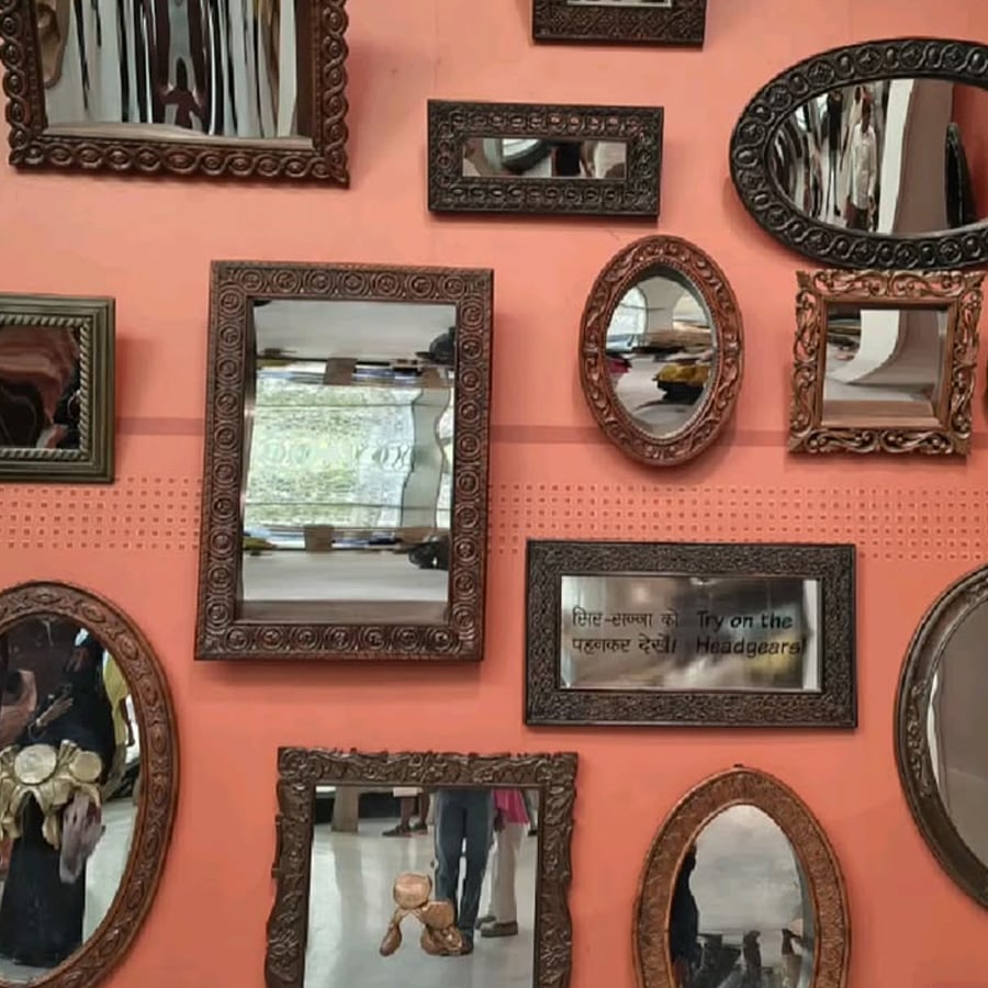 Several ornate mirrors of various shapes and sizes are mounted on a peach-colored wall, reflecting distorted images of the room and people facing them. One mirror has a sign that says, “Try on the Headgears.”