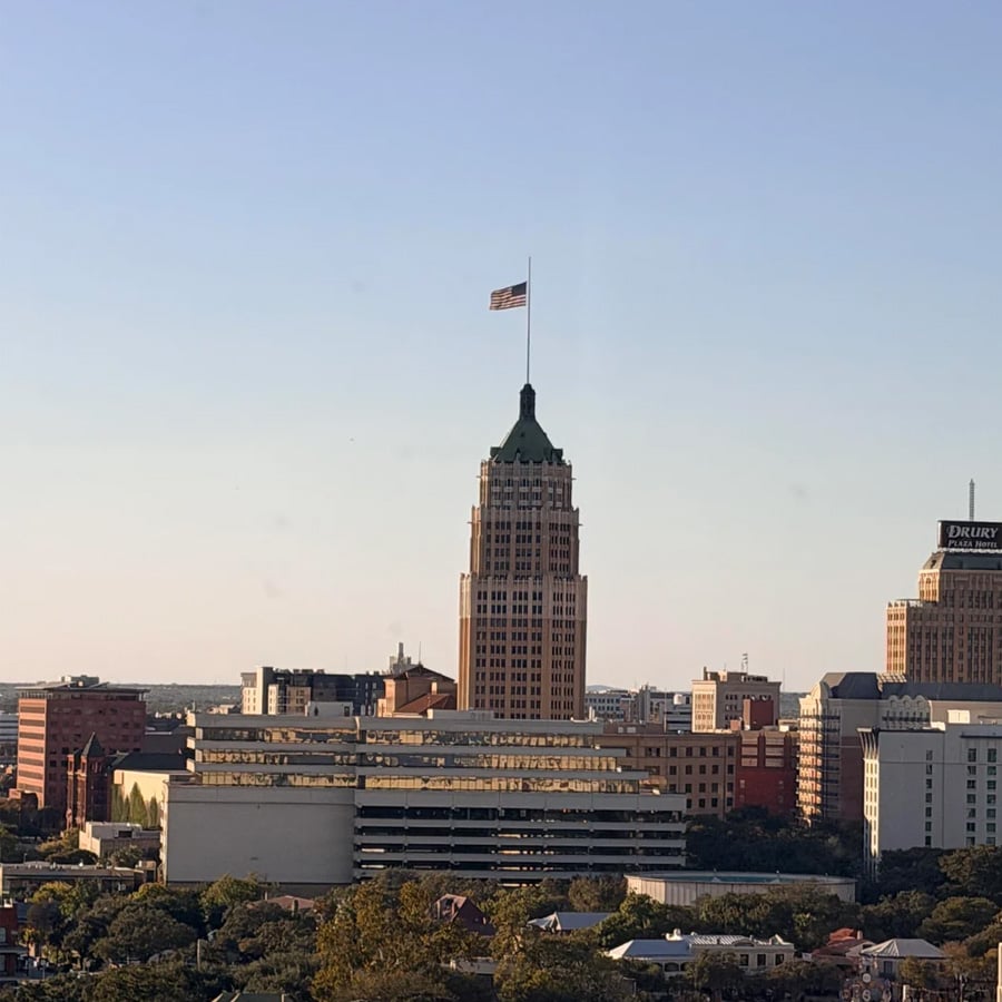 A city skyline with a tall building featuring a green peak and a US flag on top, surrounded by other buildings under a clear blue sky. Trees are visible in the foreground.