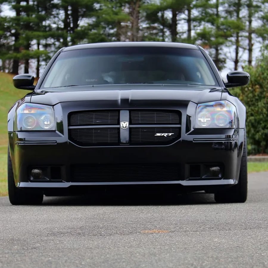 A black Dodge Magnum SRT car is parked on a paved road, surrounded by green grass and pine trees in the background.