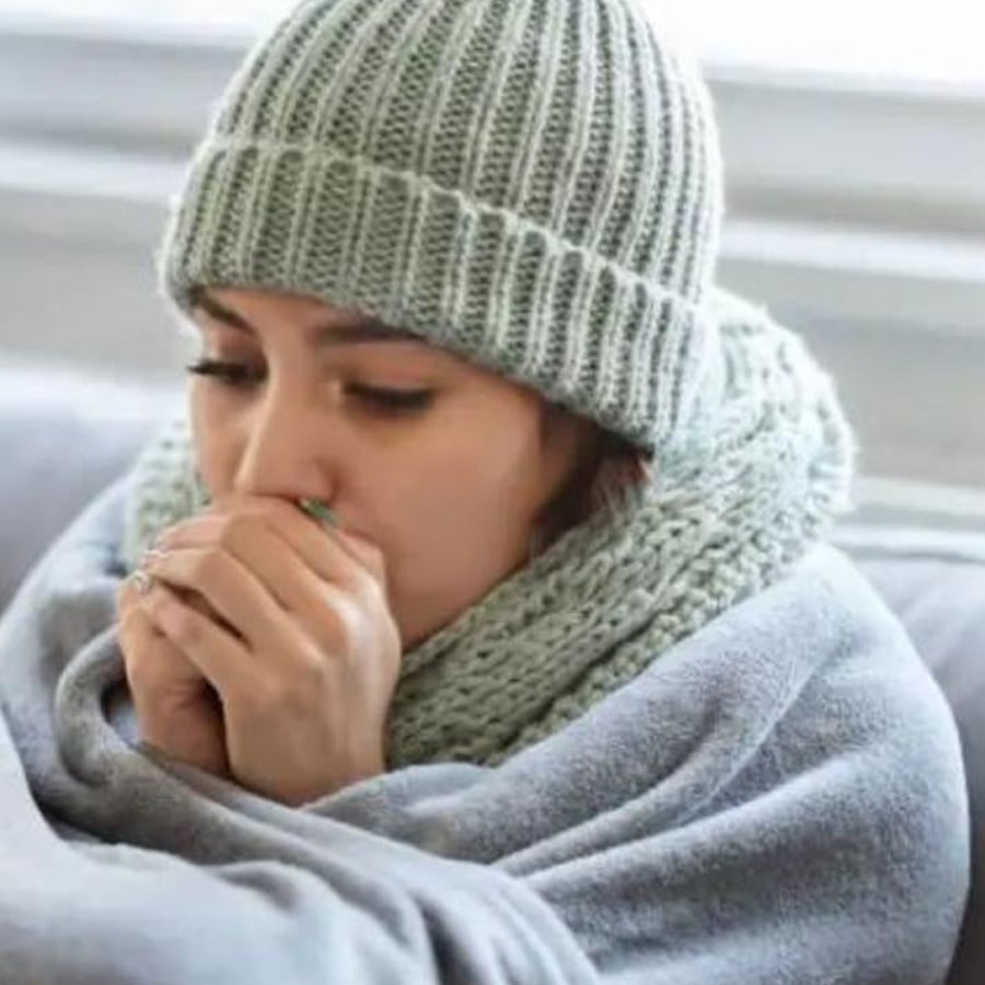 A person bundled in a gray knit hat and scarf sits on a couch, wrapped in a blanket, holding their hands to their mouth as if feeling cold or unwell.
