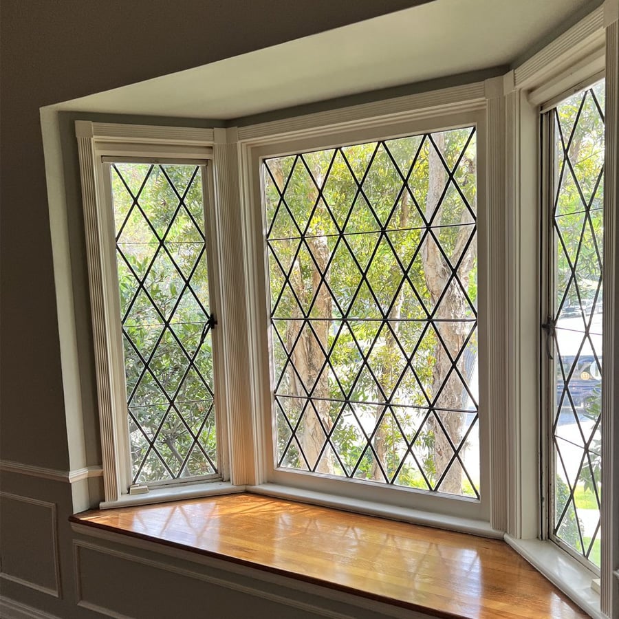 A bay window with three diamond-patterned glass panes, white trim, and a wooden windowsill. Green trees and sunlight are visible outside.