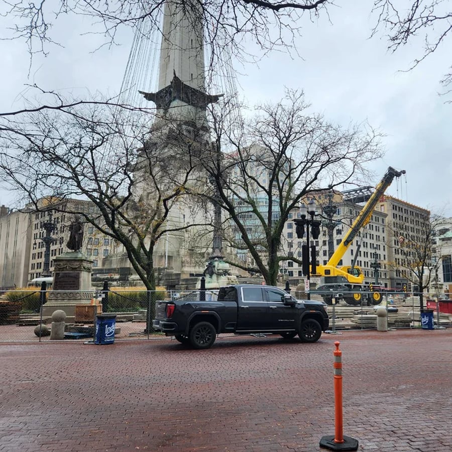 A black pickup truck is parked on a brick street near a fenced construction area with a yellow crane, leafless trees, and historic buildings in the background.