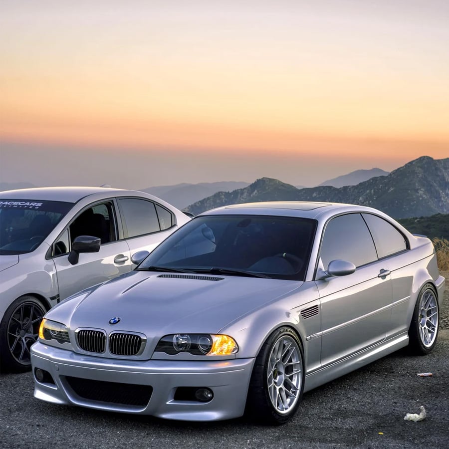 A silver BMW sports car is parked on a mountain road at sunset, with another white car partially visible beside it. The background features mountains and a colorful sky.