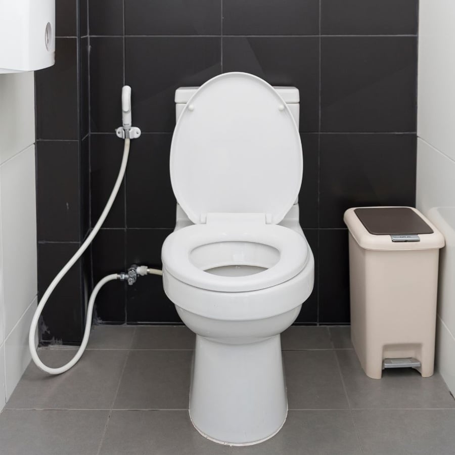 A white toilet with the lid open is next to a handheld bidet spray and a beige trash bin in a bathroom with dark tiled walls and gray flooring.