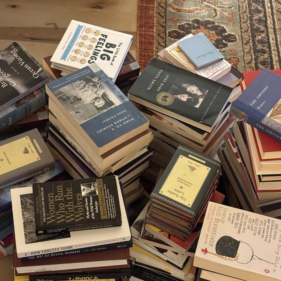 Several piles of assorted books and journals are stacked chaotically on a wooden floor next to a patterned rug. The book covers display various colors, sizes, and titles.