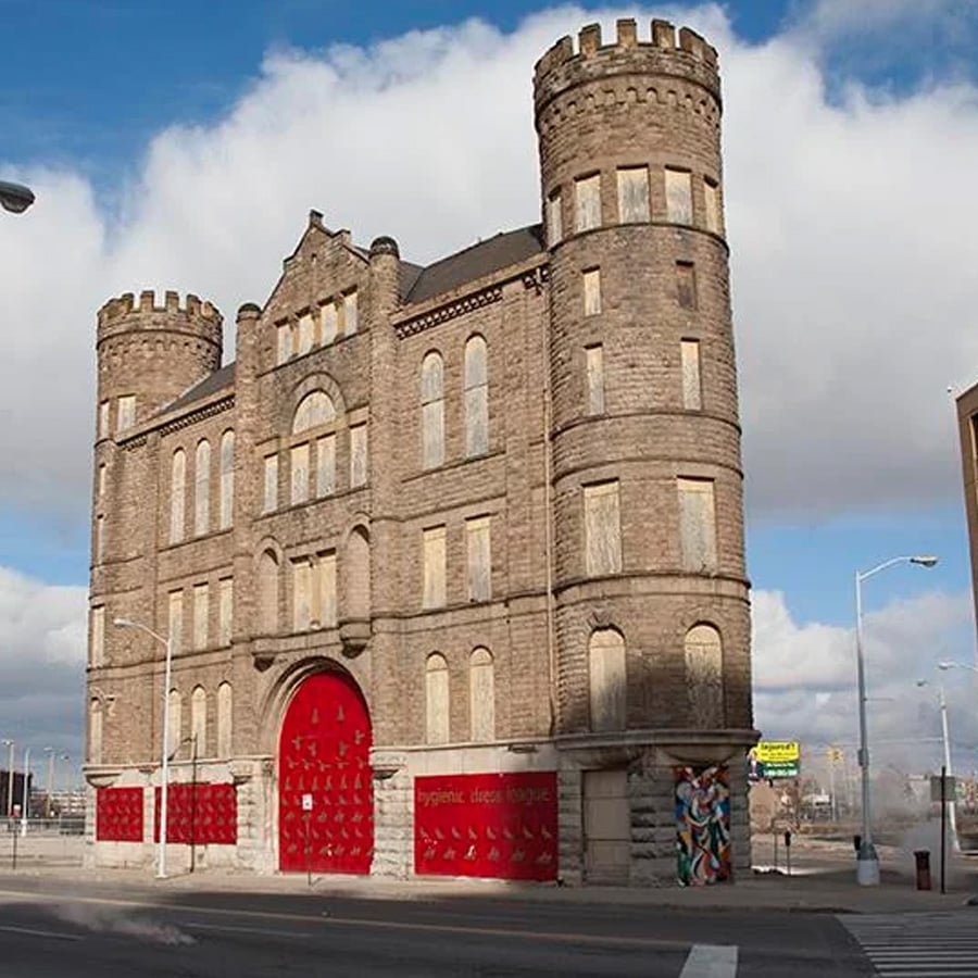 A historic stone building with round towers and large red doors, standing on a city street corner under a cloudy sky. Colorful street art is visible on the side wall near the entrance.