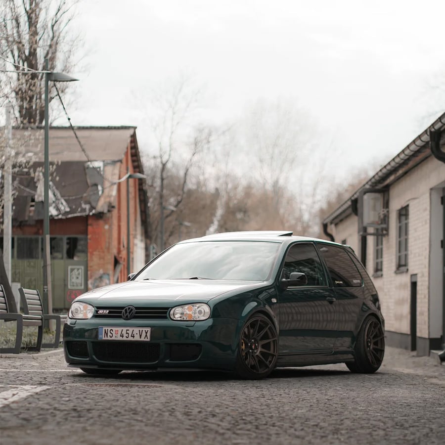 A dark green Volkswagen Golf is parked on a cobblestone street near old brick and white buildings under a cloudy sky.