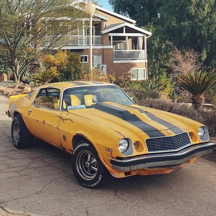 A yellow vintage muscle car with black racing stripes is parked on a street in front of a house surrounded by trees and plants.