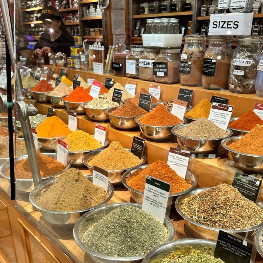 Metal bowls filled with colorful spices are displayed on a wooden counter in a shop, each labeled with a name and description. Jars of spices and herbs line shelves in the background, with a person partially visible behind the counter.