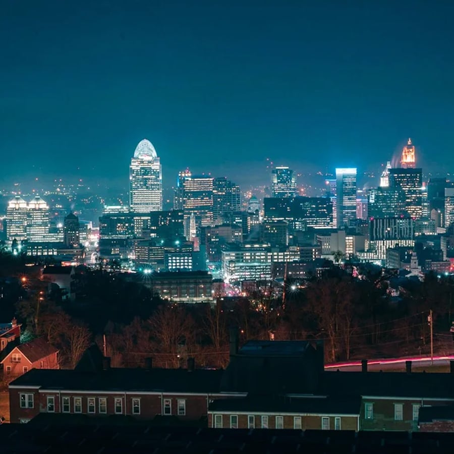 Nighttime cityscape with illuminated skyscrapers, including a distinctive domed building, against a dark sky. The city lights reflect off the buildings, while residential rooftops are visible in the foreground.