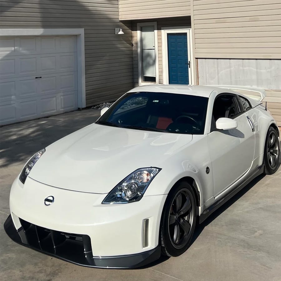 A white Nissan 350Z sports car with a large rear wing is parked on a concrete driveway in front of a beige house with a blue door and white garage.