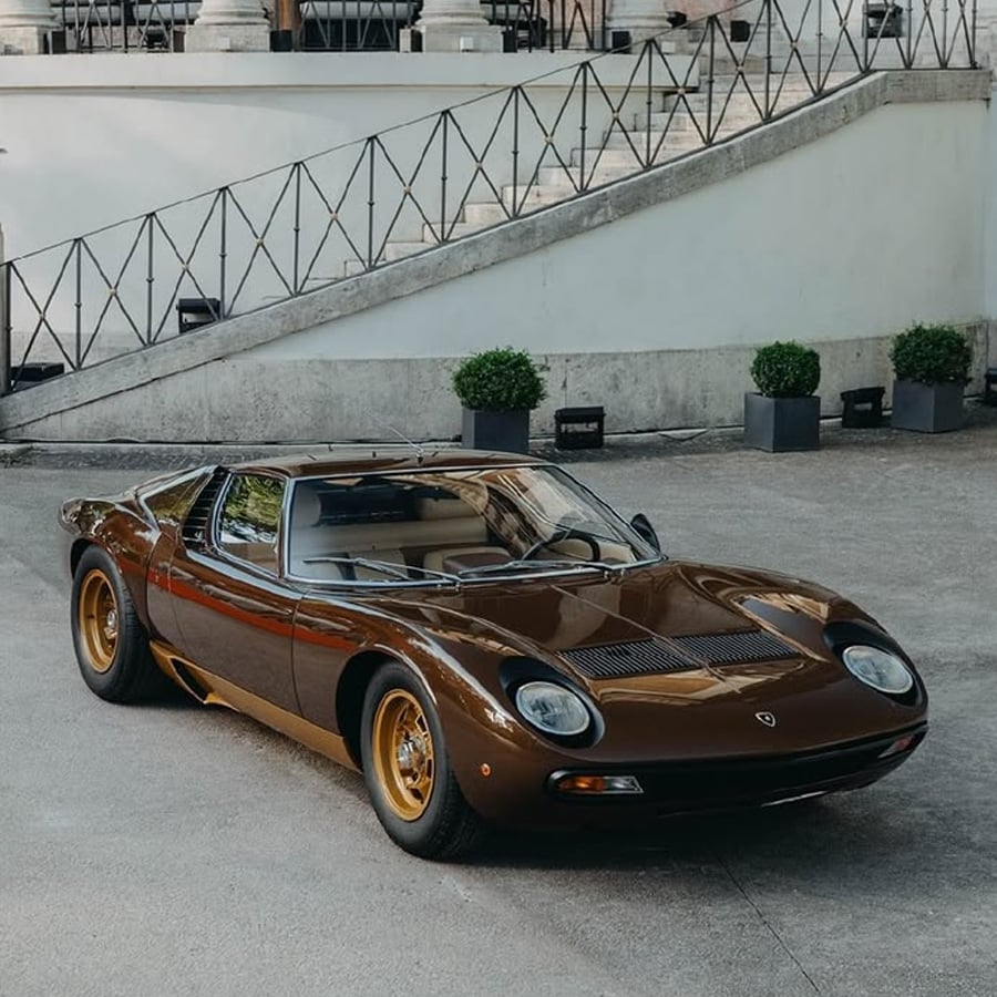 A classic brown Lamborghini Miura with gold wheels is parked on a paved driveway near a white staircase and potted plants.