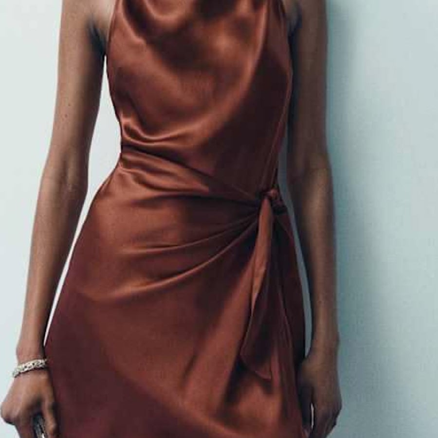 A person wearing a sleeveless, rust-colored satin dress with a waist tie stands against a light gray wall. Only the torso and upper arms are visible.