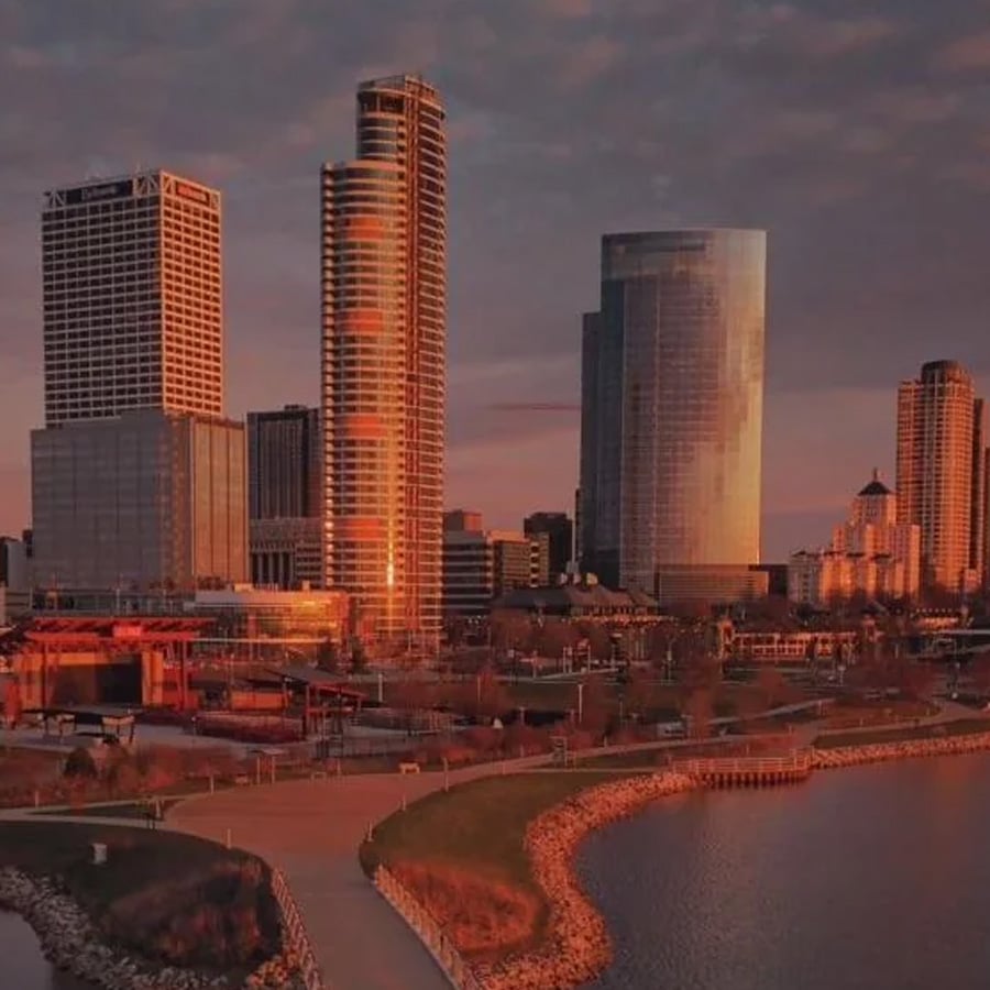 A city skyline at sunset with tall buildings reflecting warm light, a waterfront promenade, and a curved shoreline lined with rocks in the foreground.